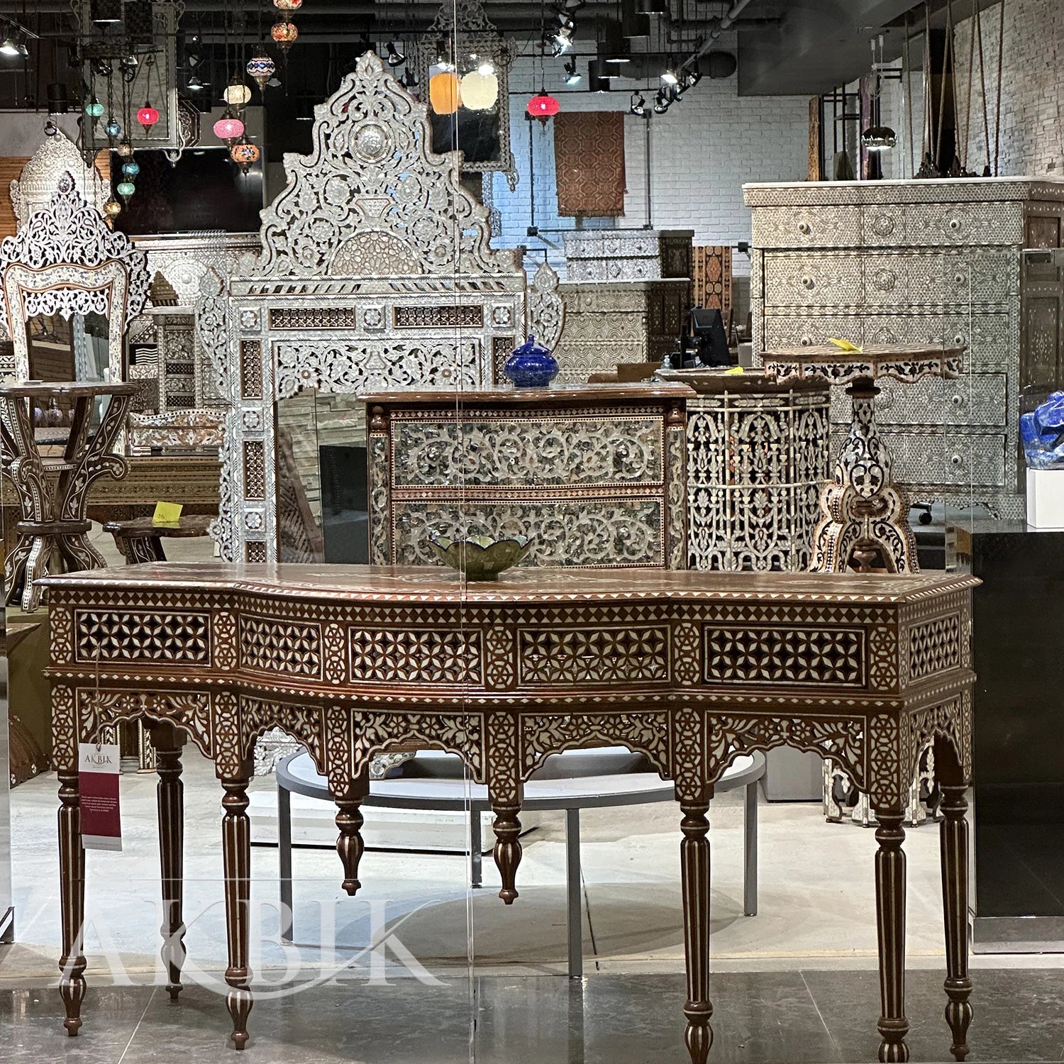 An ornate console table with intricate hand-inlaid mother-of-pearl designs on a solid teak wood structure, displayed in a furniture store setting by Akbik Furniture & Design