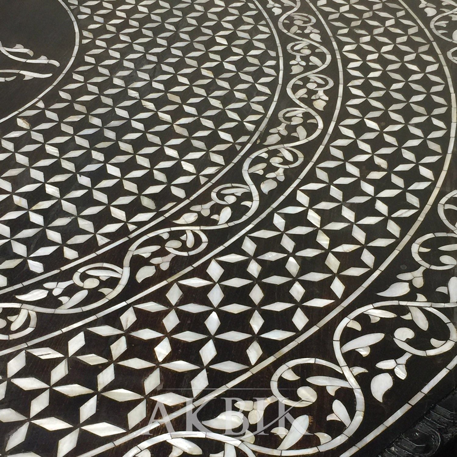 A round dining table with black and white mother of pearl inlay and hand-carved floral and geometric patterns on the pedestal base, displayed in a showroom setting with decorative items around it.