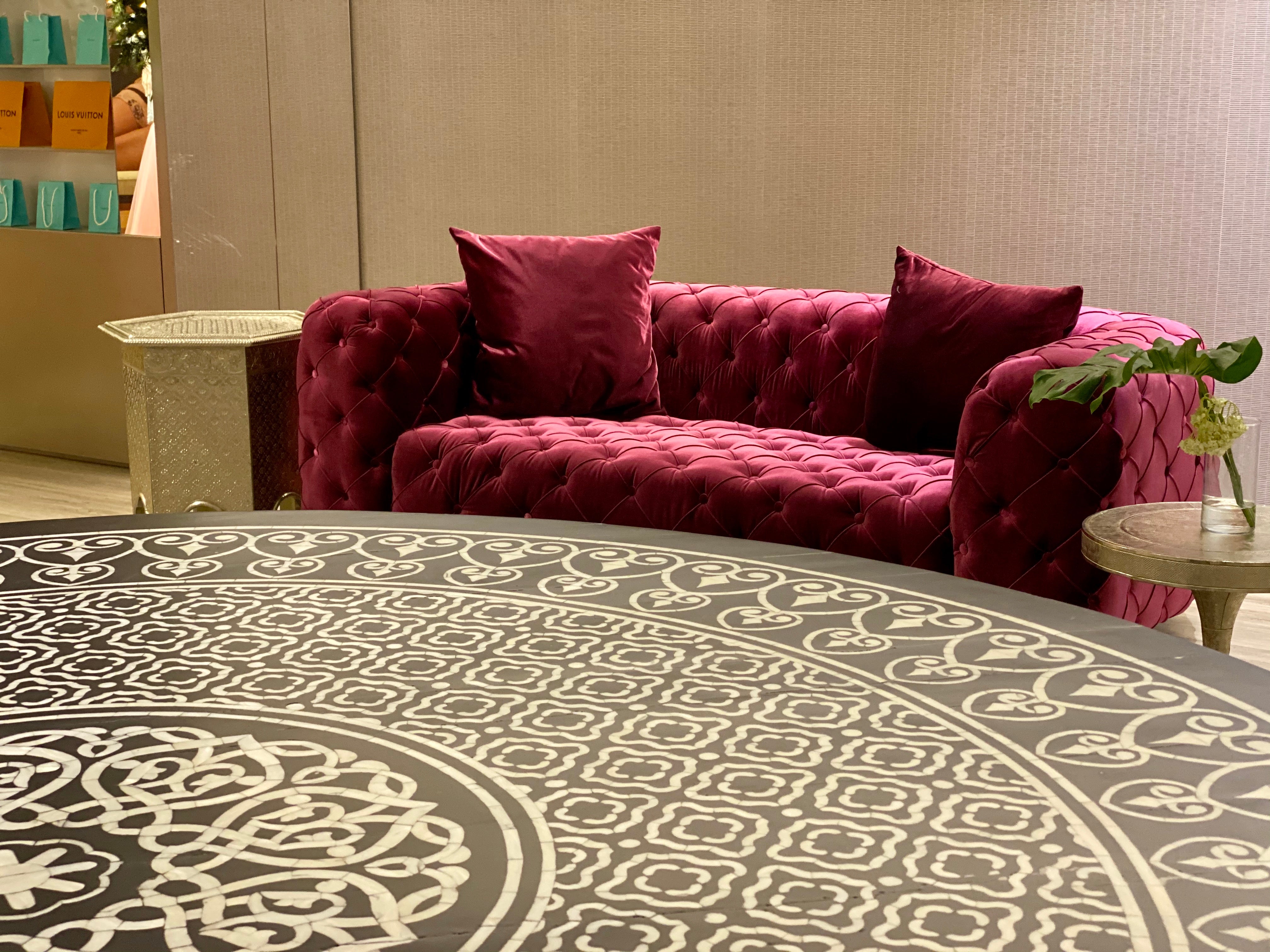 Maroon tufted sofa with decorative pillows in a room with patterned rug and wall.
