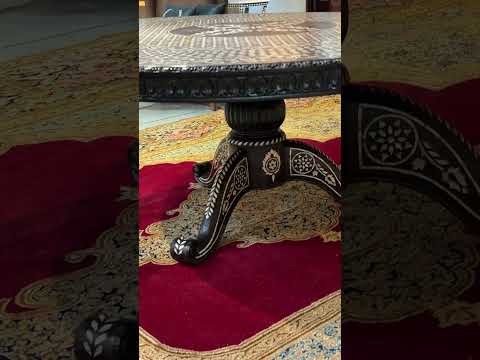 A round dining table with black and white mother of pearl inlay and hand-carved floral and geometric patterns on the pedestal base, displayed in a showroom setting with decorative items around it.