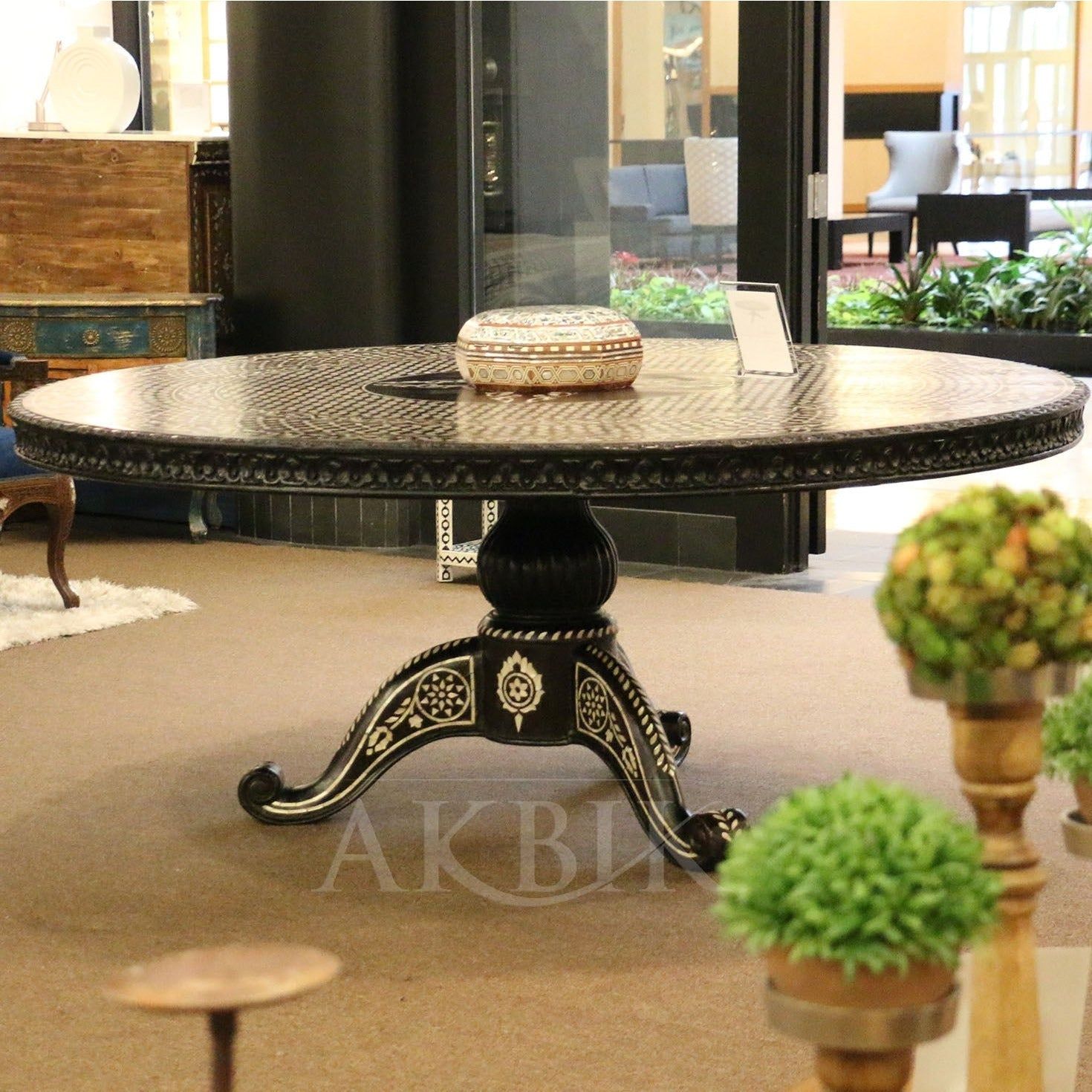 A round dining table with black and white mother of pearl inlay and hand-carved floral and geometric patterns on the pedestal base, displayed in a showroom setting with decorative items around it.